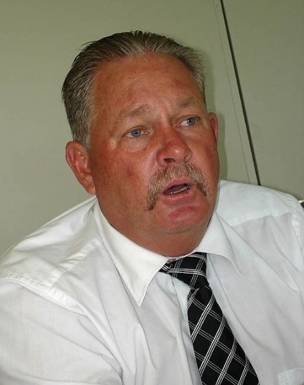 Steve Rixon with his mouth open, has white hair, and a mustache wearing a white polo long sleeve with black necktie