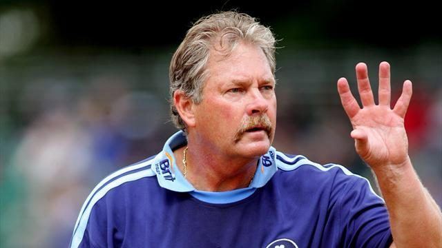 Steve Rixon seriously looking at someone with his left open hand, he has white hair, wearing a silver necklace and a blue jersey