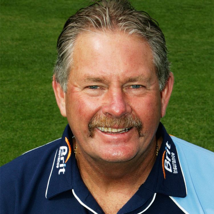 Steve Rixon smiling with his white hair inside on a football field, he is wearing a necklace and a blue jersey