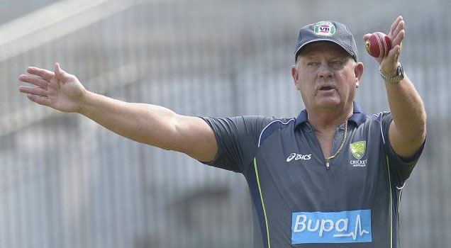 Steve Rixon talking with his L arms position while holding a red ball in his left hand, he is wearing a gray cap and gray polo jersey.