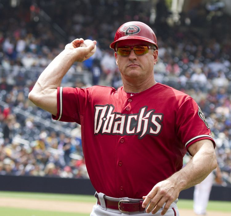 Steve Sax seriously looking afar while  throwing a ball in his left hand, wearing a red cap, sunglasses, a red jersey with “Dbacks” on it, and a red belt on a white pants