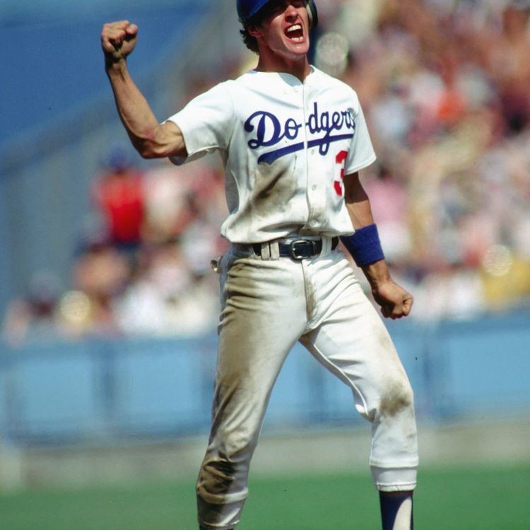 Steve Sax shouting while raising his right fist and a fist on his left hand during the baseball game (1981–88) under team Los Angeles Dodgers.  He has black hair wearing a blue cap, blue armband, black socks white jersey with “dodgers” on it, and a black belt on white pants