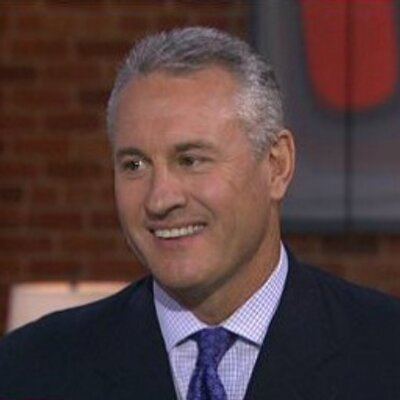 Steve Sax smiling in an interview, he has white hair, wearing a stripe purple polo long sleeve with a violet necktie under a black coat