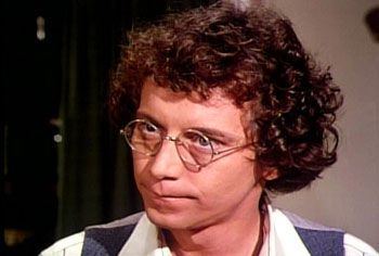 Steve Tracy with a serious face, with curly hair, and wearing eyeglasses in a scene from the television series Little House on the Prairie in the early 1980s.