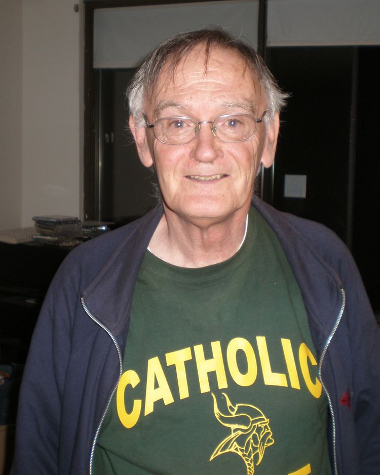 Steve Tracy smiling, with white hair, wearing eyeglasses, a blue jacket, and a green shirt.