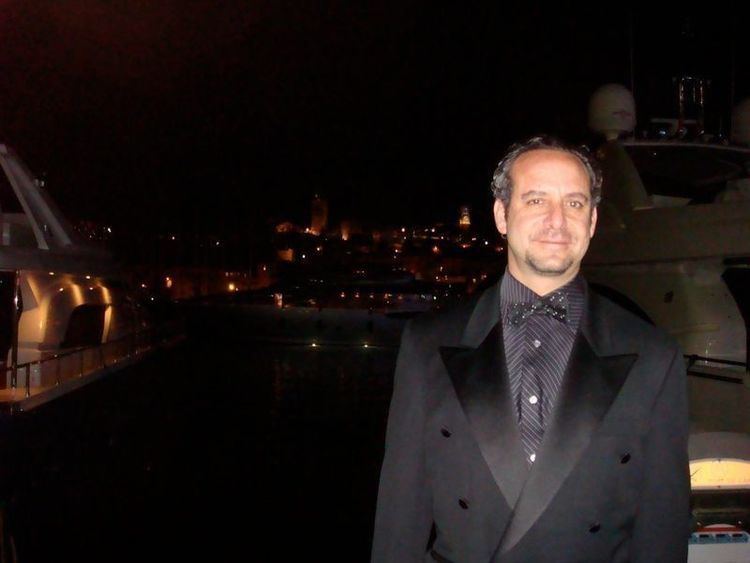 Steven G. Kaplan with a tight-lipped smile, beard, and yacht and orange lights behind him while wearing a dark blue striped long sleeve under a black polka dot bow tie, and black coat