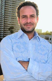 Steven G. Kaplan with a tight-lipped smile, mustache, and beard while his arms cross and wearing a light blue long sleeve