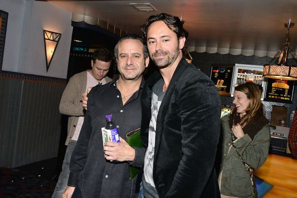 Steven G. Kaplan and James Wirt are smiling with a man and woman behind them while Steven is holding a bottle and green clutch bag at the Billy Bates LA Premiere at Los Feliz 3 Cinemas in Los Angeles, California. Steven is wearing a black long sleeve while James is wearing a gray shirt under a black coat