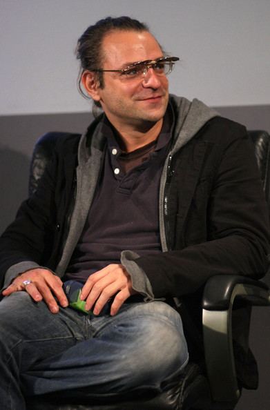Steven Martini sitting down and looking at something while wearing a black jacket over a black shirt and some eyeglasses.