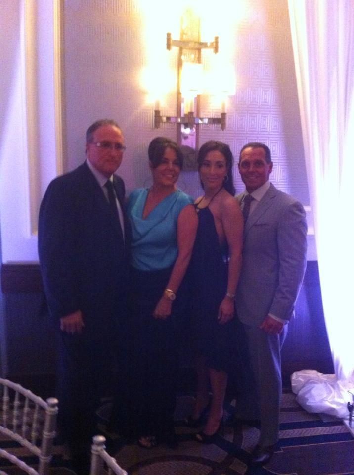 Steven Mazzone with his wife and with the other couple smiling. Steven wearing a gray suit and pants, white long sleeves, and a tie while his wife wearing a black sexy dress.