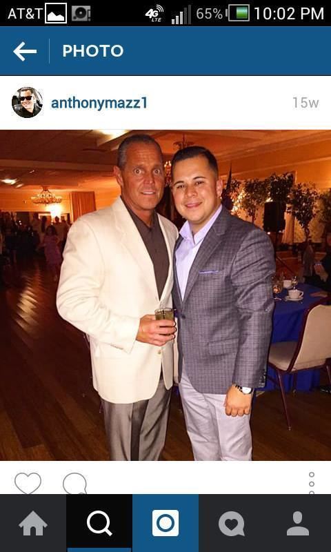 Steven and Anthony Mazzone are smiling. Steven wearing a white coat over a brown shirt, and gray pants while Anthony wearing a gray coat over white long sleeves, and khaki pants.