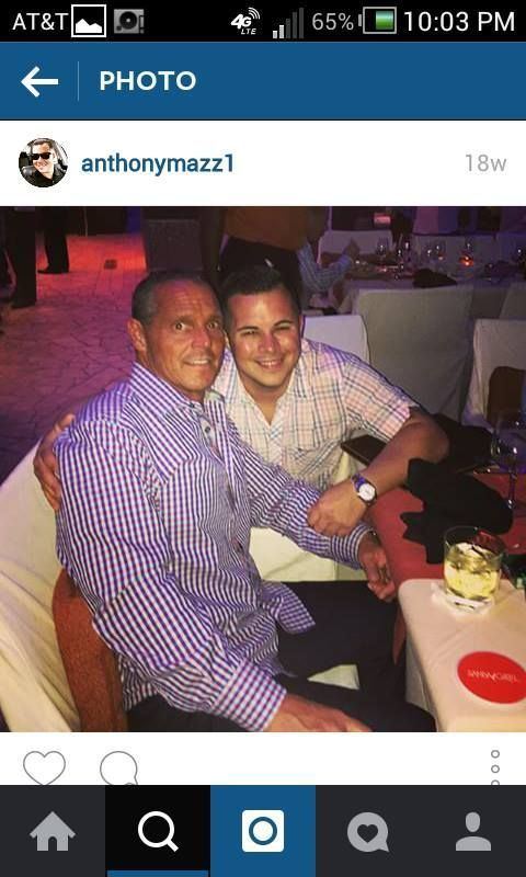 Steven and Anthony Mazzone are smiling while sitting on a couch. Steven wearing blue checkered long sleeves and black pants while Anthony wearing an off-white checkered polo shirt and a watch.