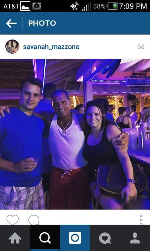 Daniel, Steven, and Savannah Mazzone are smiling inside a bar. Daniel wearing a blue shirt and checkered shorts, Steven wearing a white sleeveless shirt and red shorts while Savannah wearing a black sleeveless top and black shorts.