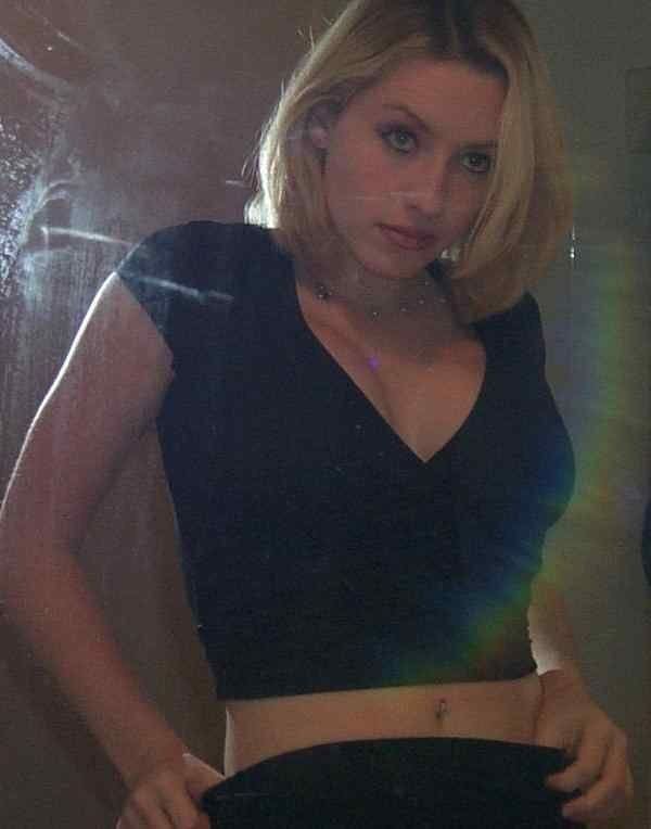 In a room with a large mirror, Stevie Case is serious, standing holding her pants on her waist, has blond hair and a navel piercing, wearing earring, bead necklace, a black showing cleavage shirt and a black pants.