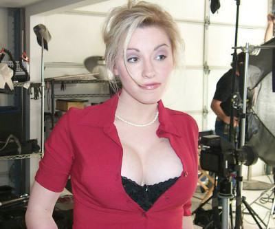 In a studio room, with white walls, lights and reflectors, Stevie case is serious, standing, has blonde hair wearing a white necklace black bra and a red showing cleavage polo shirt.