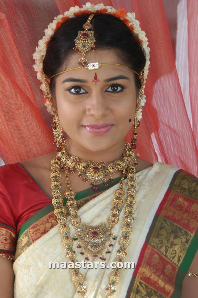 Sudeepa Pinky with a tight-lipped smile while wearing orange, cream, and green dress, gold necklace with stones, earrings, and head dress