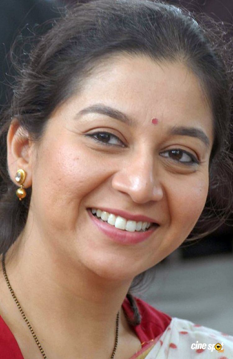 Sudha Rani smiles while wearing earrings and a red and white shirt