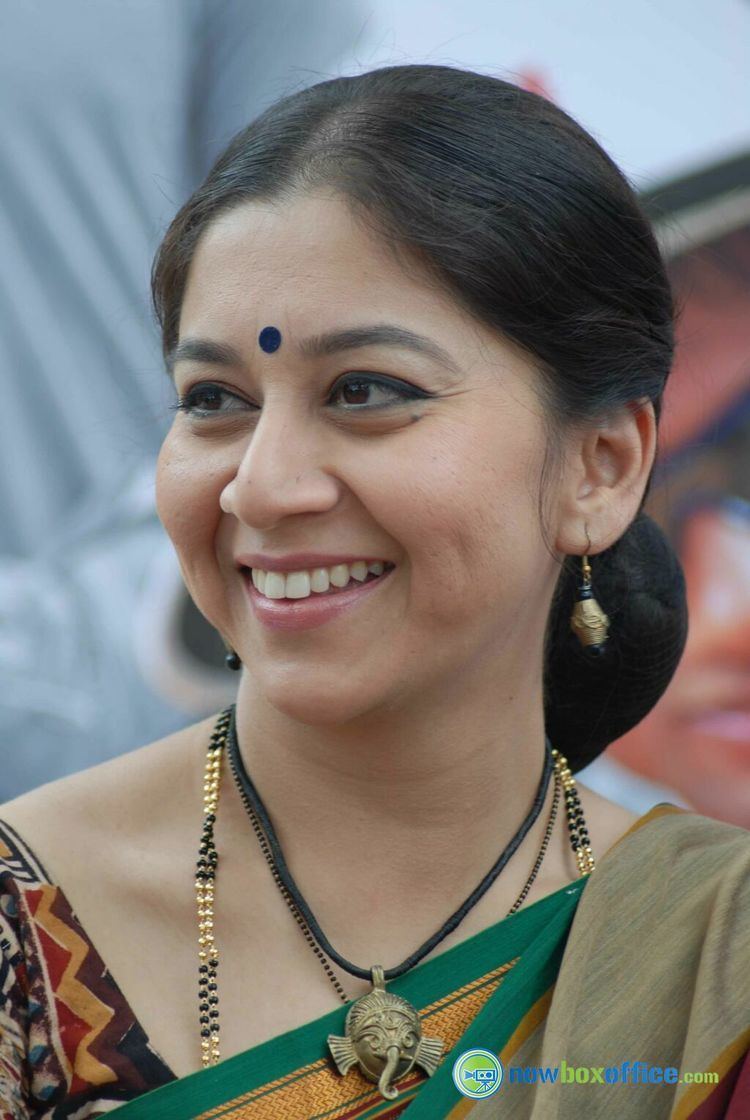 Sudha Rani smiles while wearing a printed shirt, necklaces, and pair of earrings