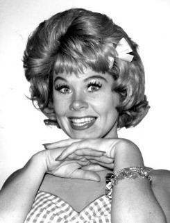 Sue Ane Langdon with a big smile and ribbon on her short wavy hair while hands on her chin and she is wearing a bracelet and a checkered top with a sweetheart neckline