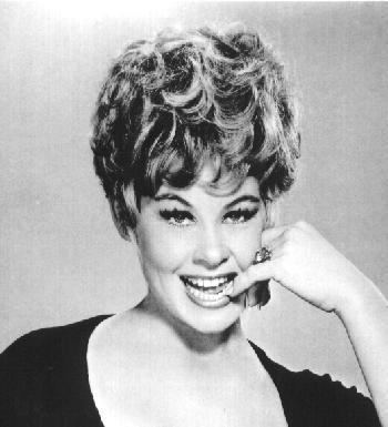 Sue Ane Langdon with a big smile, short wavy hair, and her pinky finger is on her mouth. Sue is wearing a ring and blouse