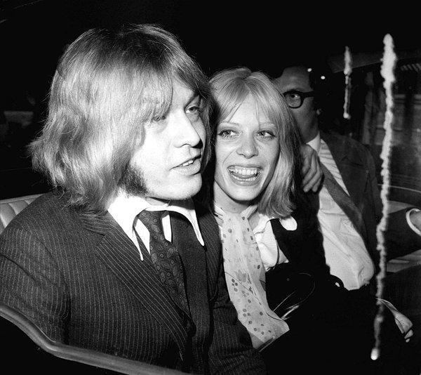 Suki Potier smiling and going with Brian Jones while wearing a white dress along with a white neckerchief.