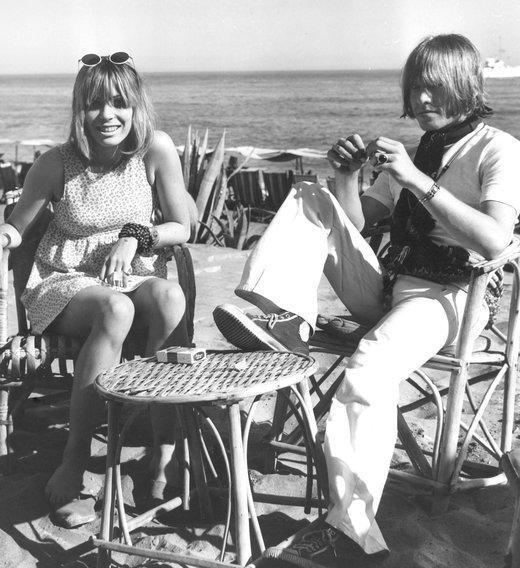 Suki Potier sitting down at the beach with Brian Jones and wearing a white spotted sleeveless dress.