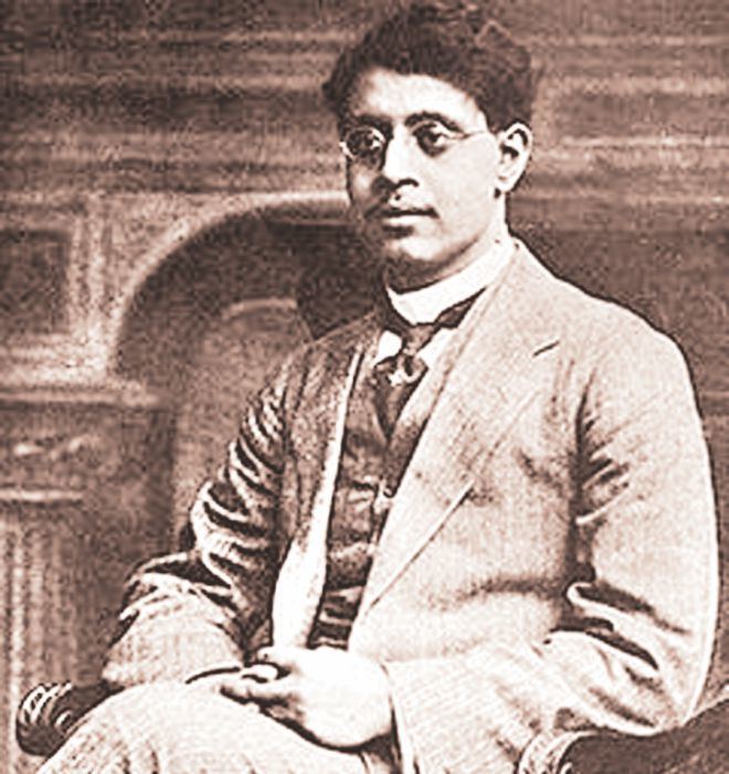 Sukumar Ray sitting on the chair while wearing eyeglasses, striped coat, vest, long sleeves, necktie, and striped pants
