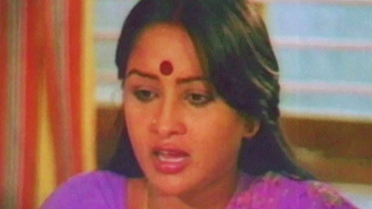 Sulakshana is talking while looking at something with a Bindi on her forehead and nose pin and wearing a purple saree dress.