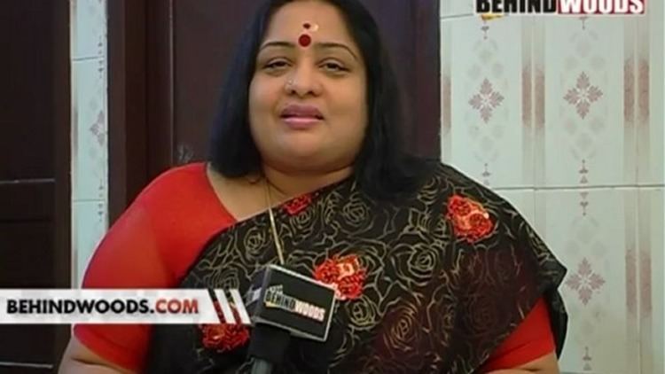 Sulakshana talking in a microphone in her front, with a Bindi on her forehead, and a nose pin,  wearing a necklace and a red and black saree dress.