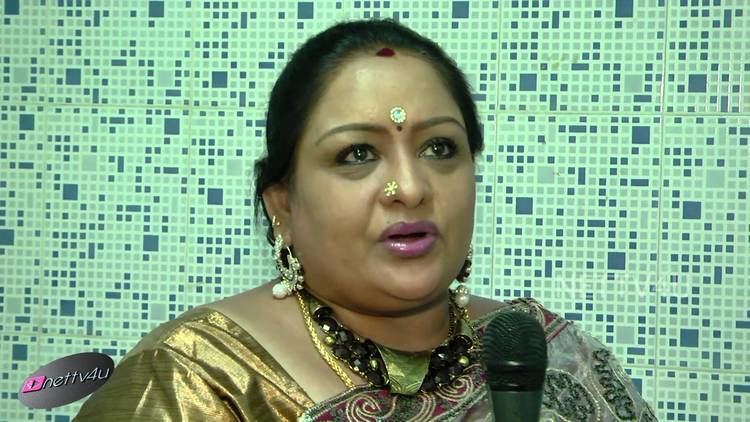 Sulakshana is talking in a microphone in her front, with a Bindi on her forehead, and a nose pin, wearing earrings, necklaces, and a multi-colored saree dress.