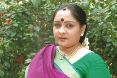 Sulakshana smiling, with a Bindi on her forehead, a flowers & vermilion in her hair, and a nose pin, wearing earrings, necklaces, wearing a green & purple saree dress