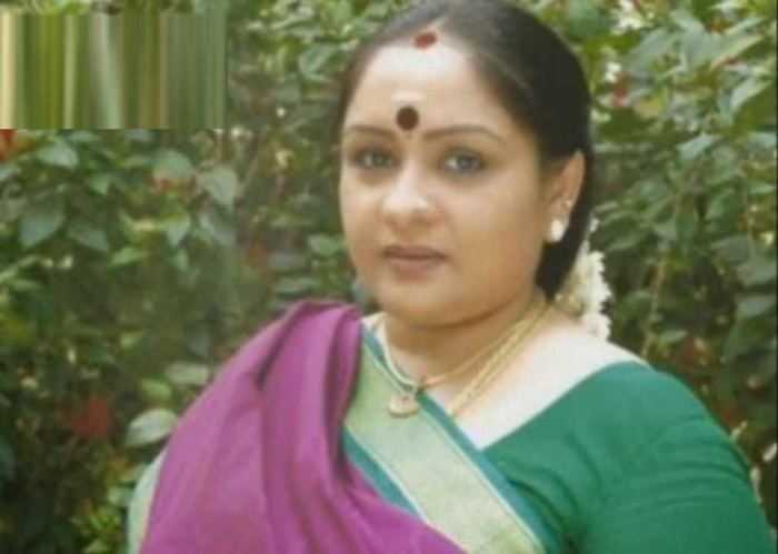 Sulakshana smiling, with a Bindi on her forehead, a flowers & vermilion in her hair, and a nose pin, wearing earrings, necklaces, wearing a green & purple saree dress