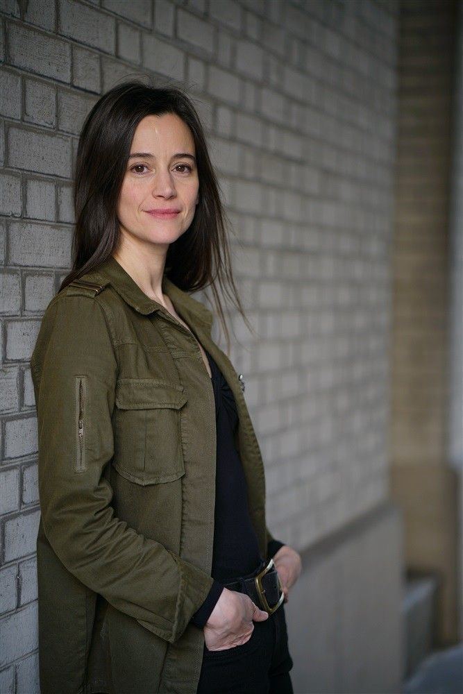 Suliane Brahim smiling while leaning on the wall and hands on her pockets, wearing a shade of green coat, black shirt, black pants, and black belt.