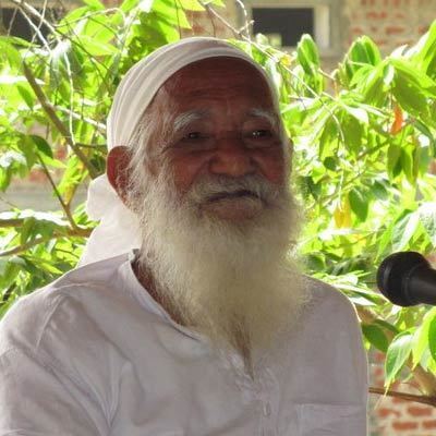 Sunderlal Bahuguna with a white long mustache and beard, wearing a white bandana and a white shirt with a microphone in front of him.