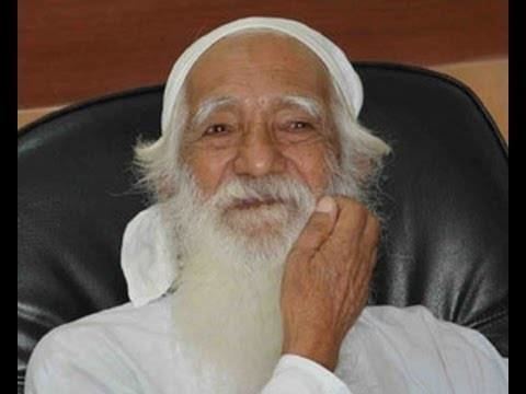 Sunderlal Bahuguna sitting on a black chair, with a white long mustache and beard, wearing a white bandana and a white shirt while scratching his face.
