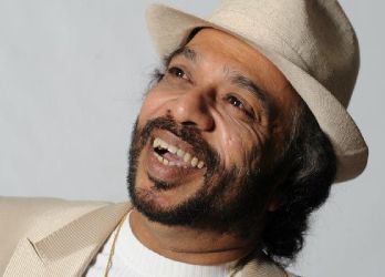 Sunil Perera smiling with thick facial hair and wearing a white hat and suit.