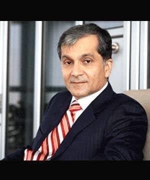 Surendra Hiranandani with a serious face while sitting on a couch, wearing a black coat over white long sleeves, and a red striped necktie.