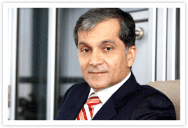 Surendra Hiranandani with a serious face, wearing a black coat over white long sleeves, and a red striped necktie.