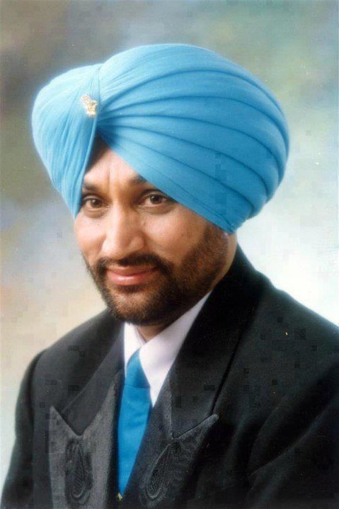 Surjit Bindrakhia smiling while looking into something and wearing a white sleeve, black suit, blue necktie and blue turban