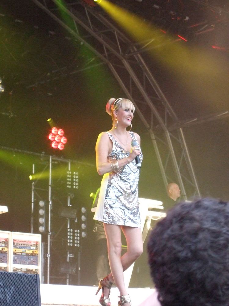 Susan Ann Sulley smiling while performing and wearing a silver sleeveless dress, heels, and some pieces of jewelry