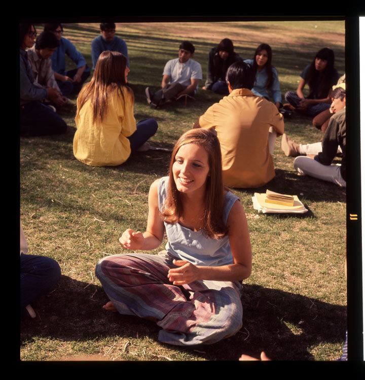 Susan Foster sitting on the ground at the set of the 1971 film 'Billy Jack' while wearing a blue sleeveless top and blue and red pants