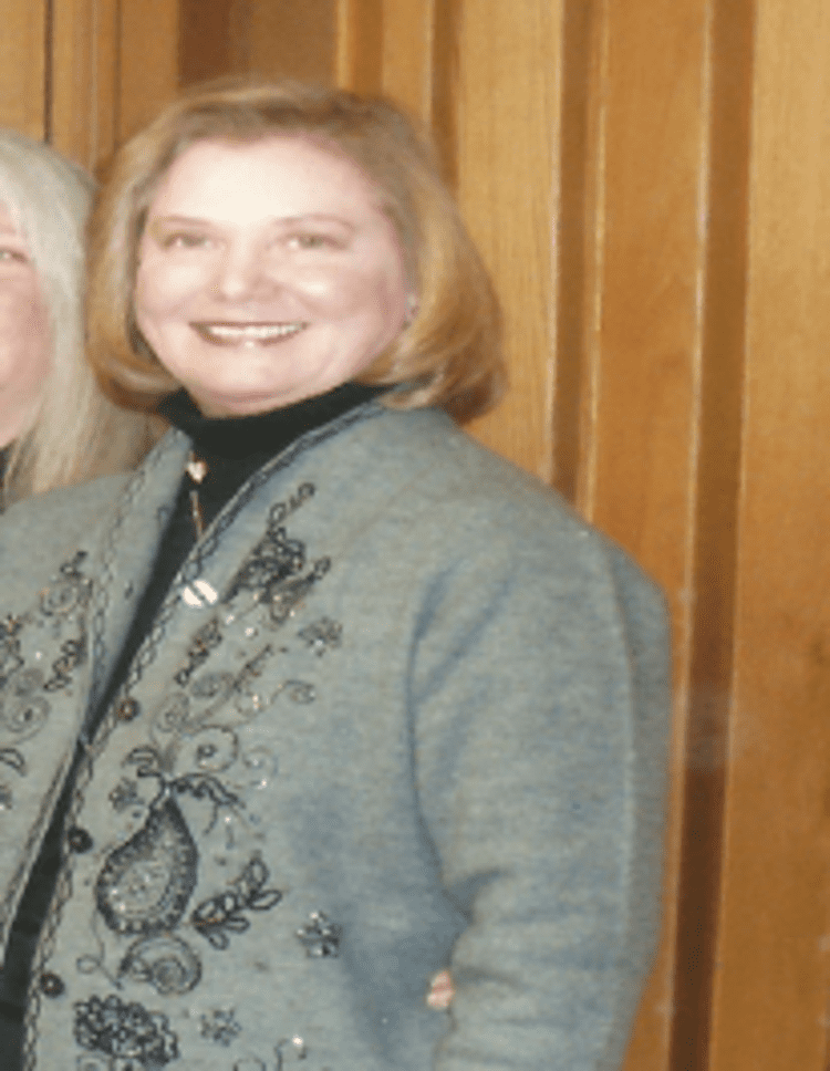 Susan Headley smiling beside a girl with blonde hair. Susan with short blonde hair, wearing a necklace and a gray coat with a flower design over a black turtleneck top.