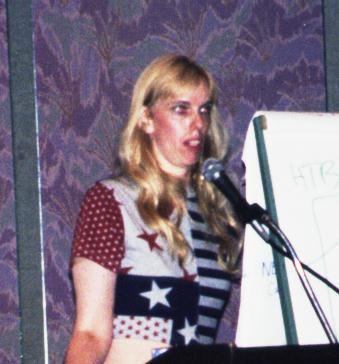 Susan Headley aka “Susy Thunder” is talking in front of a microphone, with wavy long blonde hair and wearing a multi-colored dress with stripes and stars designs.