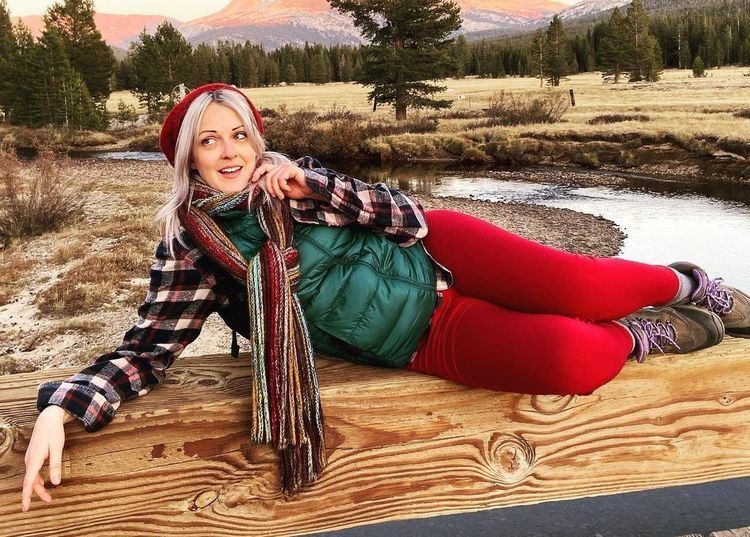 Susan Slaughter smiling and lying on a wood in a sideway position while wearing a green coat, checkered sleeve, red pants, brown shoes, shawl, and a red cap