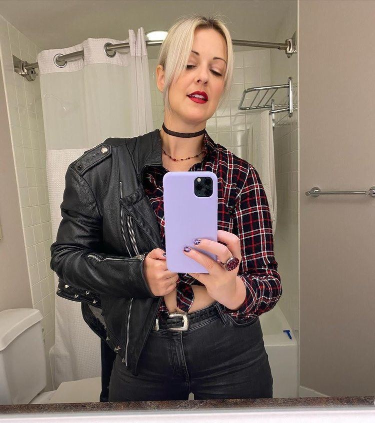 Susan Slaughter taking a selfie in front of the mirror while wearing a checkered blouse, black leather jacket, and black pants