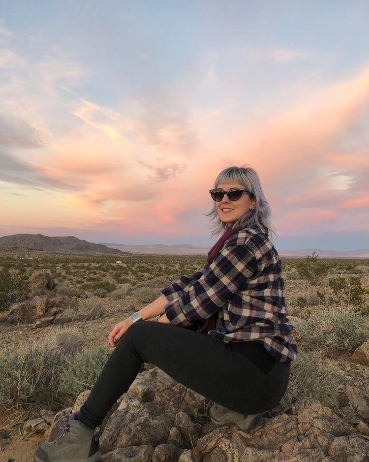 Susan Slaughter smiling while sitting on a rock in a side view while wearing a checkered shirt, black pants, and green boots