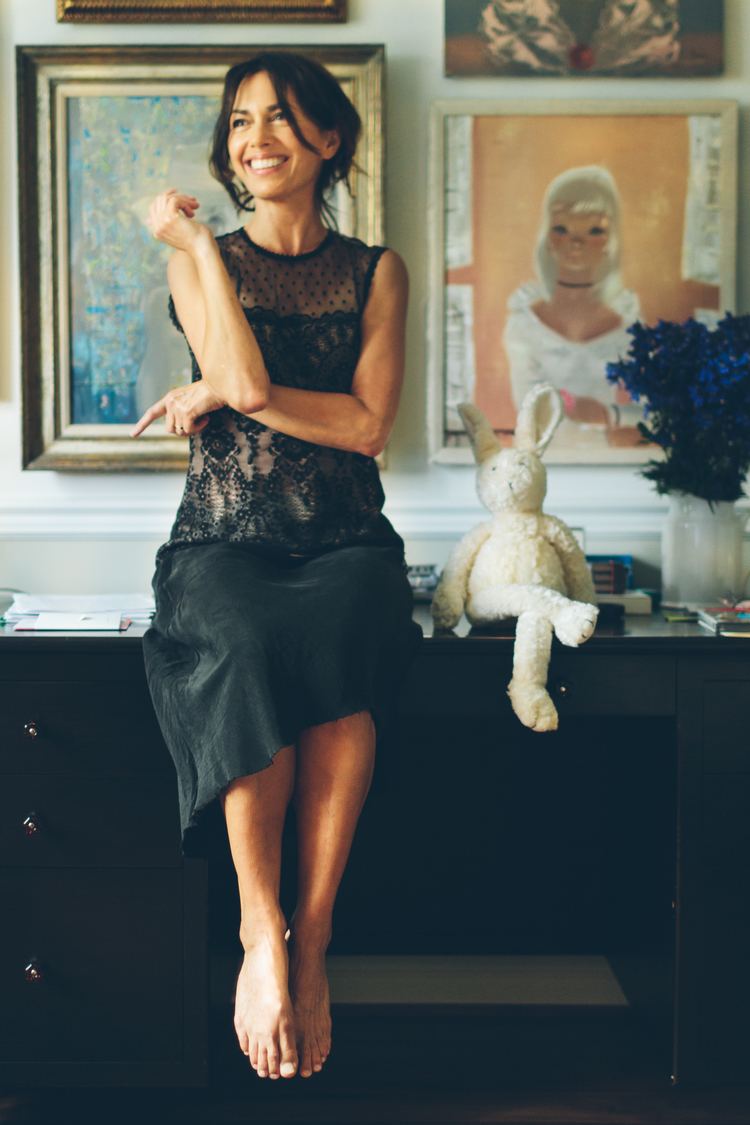 Susanna Hoffs is smiling while looking afar beside a bunny with flowerpot & paintings in the background and wearing a black lace dress.