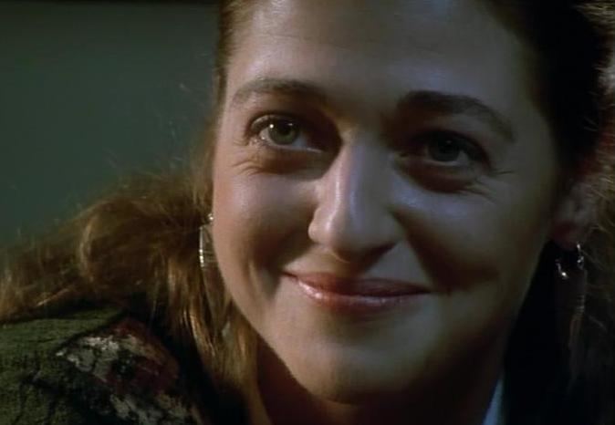 Susannah Corbett as Ellie Pascoe, with a tight-lipped smile while wearing earrings and a green and white blouse in a scene from the 1996 tv series, Dalziel and Pascoe