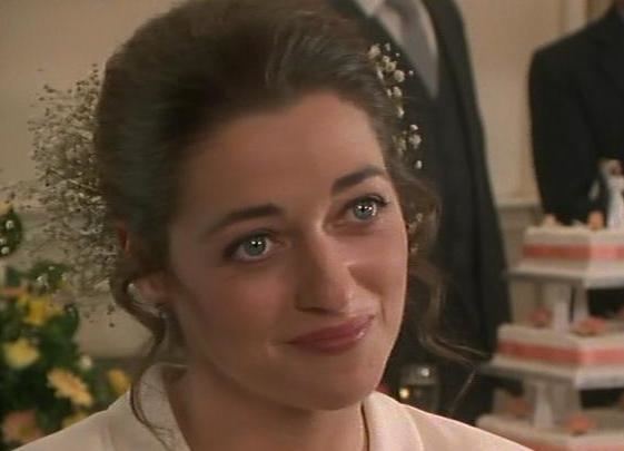 Susannah Corbett as Ellie Pascoe, with a tight-lipped smile and curly hair, while wearing a white blouse in a scene from the 1996 tv series, Dalziel and Pascoe