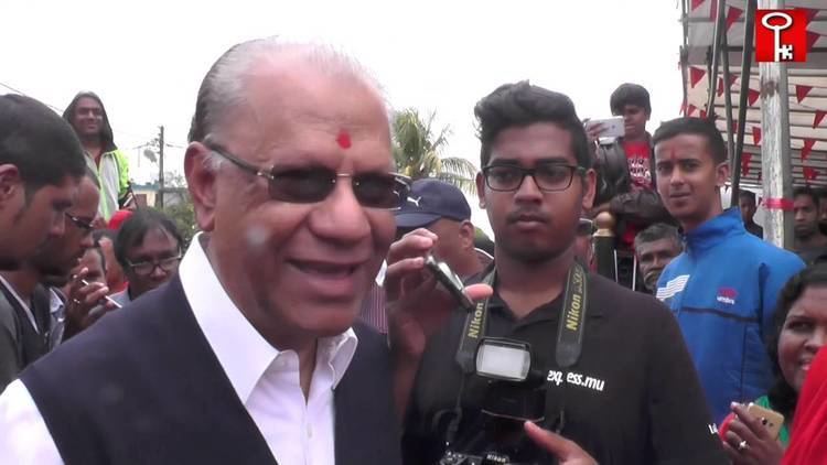 Seewoosagur Ramgoolam, husband of Sushil Ramgoolam wearing eyeglasses and a black and white suit surrounded by media.